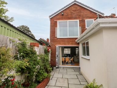 A backyard with paved flooring plants along a wooden fence and a red brick house with glass doors open to a dining area at The Headlands - Gower in The Mumbles