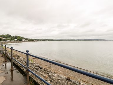 A view of a beach with a stone wall and metal railing overlooking water and distant hills at The Headlands - Gower in The Mumbles