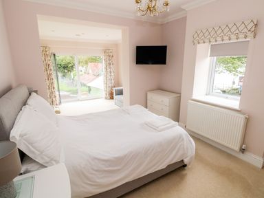 A bedroom with a double bed white bedding a chandelier a wall-mounted TV a chest of drawers and windows with floral curtains at The Headlands - Gower in The Mumbles
