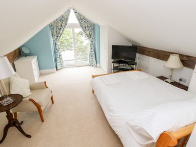 A bedroom with a bed, armchair, chest of drawers, television, and window with curtains at The Headlands - Gower in The Mumbles