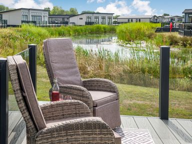 Two wicker chairs with cushions on a balcony overlooking a pond and multiple modern houses at Number 8 Lakes View in Brean