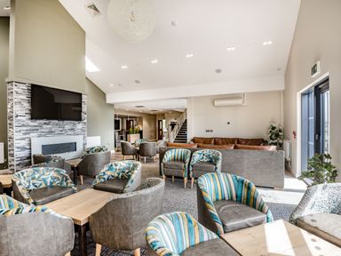 A lounge area with patterned armchairs and sofas around tables a television above a fireplace and a staircase at Number 8 Lakes View in Brean