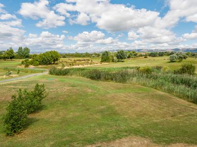 A golf course with green grass trees and a pathway under a cloudy blue sky at Number 8 Lakes View in Brean