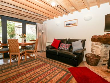 A living room with a black sofa with cushions a wooden dining table with chairs a patterned rug and a stone fireplace at Tinkley Cottage in Nympsfield near Nailsworth