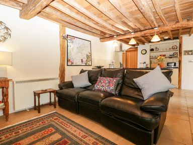 A living room with a black leather sofa with cushions a rug and a wooden beamed ceiling at Tinkley Cottage in Nympsfield near Nailsworth