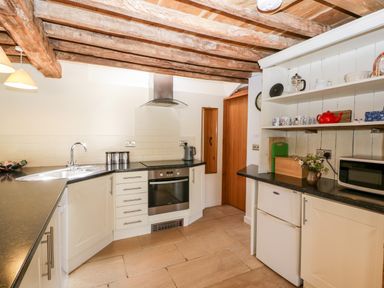 A kitchen with wooden beams on ceiling a sink oven microwave and shelves with mugs at Tinkley Cottage in Nympsfield near Nailsworth