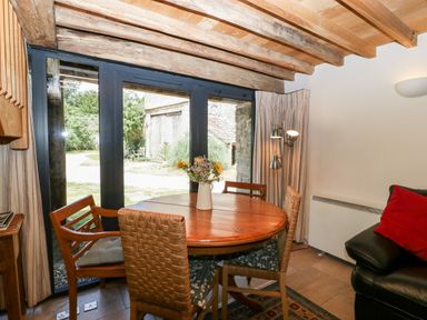A dining area with a round wooden table and chairs next to large glass doors and a black leather sofa at Tinkley Cottage in Nympsfield near Nailsworth