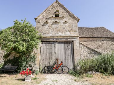 A stone building with a wooden door two bicycles a bench a chair a tree and a flower pot at Tinkley Cottage in Nympsfield near Nailsworth