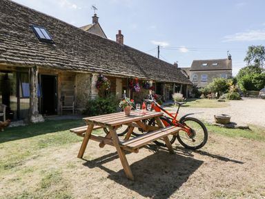 A picnic table with two wine glasses and a bottle next to a bicycle outside a stone cottage with hanging flower baskets at Tinkley Cottage in Nympsfield near Nailsworth