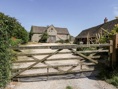 A wooden gate with a sign reading Bay Tree Farm in front of stone farm buildings and a gravel driveway at Tinkley Cottage in Nympsfield near Nailsworth