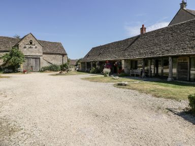 A courtyard with gravel ground between stone buildings with wooden doors and flower pots at Tinkley Cottage in Nympsfield near Nailsworth