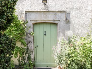 A green wooden door set in a stone frame with plants growing around it at Tinkley Cottage in Nympsfield near Nailsworth