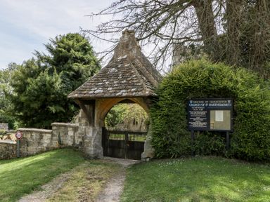 An entrance gate with a tiled roof leading to a churchyard with a sign for Church of St Bartholomew at Nympsfield near Nailsworth