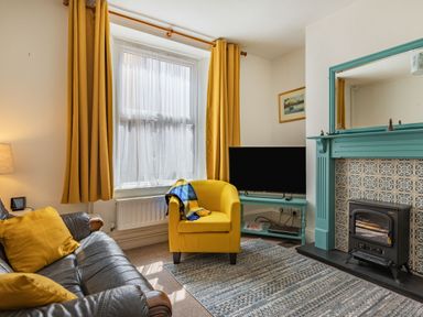 A living room with yellow curtains and armchair a black sofa and a teal fireplace at Sea Spray in Watchet
