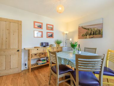 A dining room with a round table with a tablecloth and chairs next to a wooden sideboard with baskets and board games at Sea Spray in Watchet