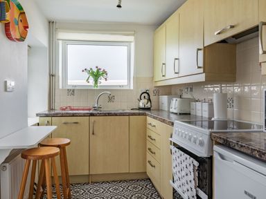 A kitchen with wooden cabinets, a stove, a sink under a window with flowers, a toaster, kettle, and two wooden stools at Sea Spray in Watchet