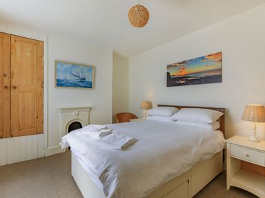 A bedroom with a double bed two bedside tables lamps a wicker chair a wooden wardrobe and maritime paintings at Sea Spray in Watchet