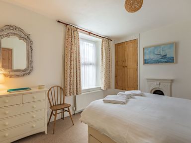 A bedroom with a bed folded towels a dresser with a mirror a wooden chair floral curtains and a painting of a ship at Sea Spray in Watchet