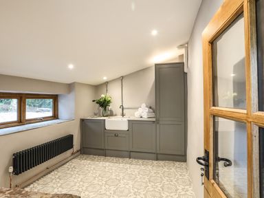 A utility room with grey cabinets a white sink folded towels a vase with flowers a black radiator and patterned floor tiles at Yr Hen Oriel in Llanrug
