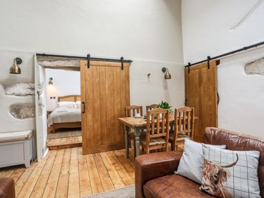 A dining area with a wooden table and chairs next to sliding wooden doors leading to a bedroom with a bed visible a leather sofa with cushions in the foreground at Yr Hen Oriel in Llanrug