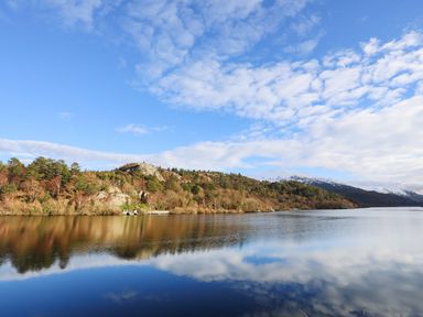 A lake with trees on the shore and snow-capped mountains in the background under a partly cloudy sky at Yr Hen Oriel in Llanrug
