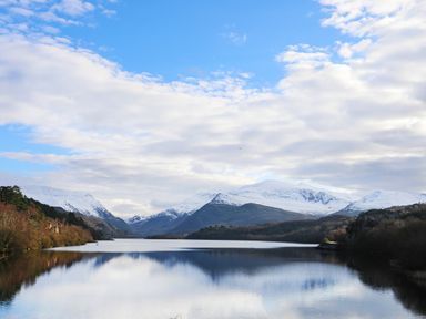 A lake with snow covered mountains and trees under a partly cloudy sky at Yr Hen Oriel in Llanrug