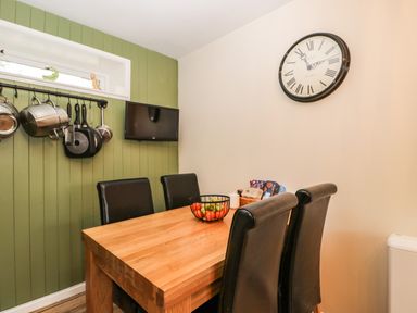 A dining area with a wooden table four black chairs a wall clock and kitchen utensils hanging on a green wall at Orchard House Cottage Malmesbury