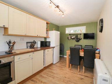 A kitchen with wooden floor cream cabinets dining table with black chairs hanging pots on green wall at Orchard House Cottage in Malmesbury