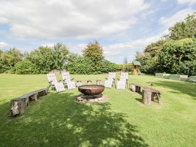 A garden with a fire pit surrounded by chairs and benches and a children's playset at Orchard House Cottage in Malmesbury