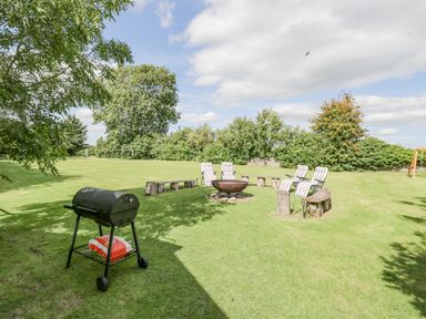 A grassy backyard with a charcoal grill a fire pit surrounded by chairs and benches and trees in the background at Orchard House Cottage Malmesbury