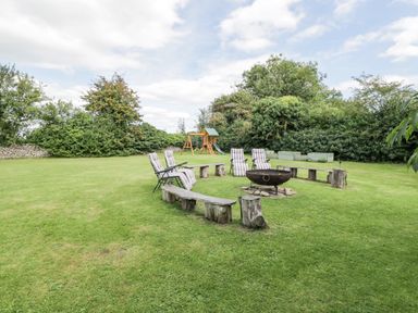 A grassy backyard with a circular wooden bench and chairs around a fire pit and a children's playset in the background at Orchard House Cottage in Malmesbury