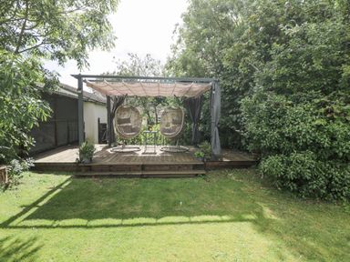 A wooden deck with two hanging chairs and a small table under a canopy in a garden at Orchard House Cottage in Malmesbury