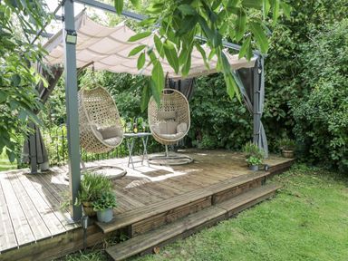 An outdoor wooden deck with two hanging wicker chairs and a small table under a canopy at Orchard House Cottage in Malmesbury