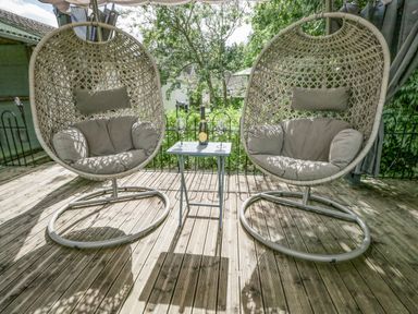 Two hanging wicker chairs with cushions and a small table with a bottle and two glasses on a wooden deck at Orchard House Cottage in Malmesbury