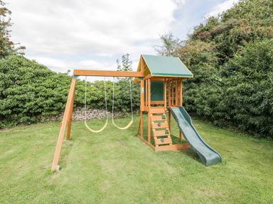A children's wooden playset with two yellow swings a green slide and a climbing wall in a grassy garden at Orchard House Cottage in Malmesbury