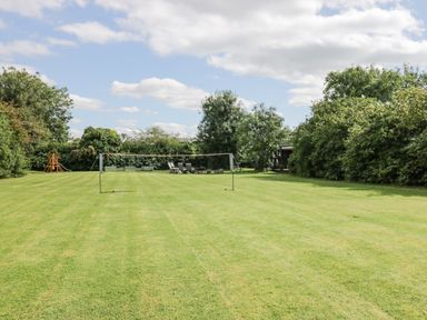 Large grassy backyard with a badminton net chairs and a children's playset surrounded by trees and bushes at Orchard House Cottage in Malmesbury