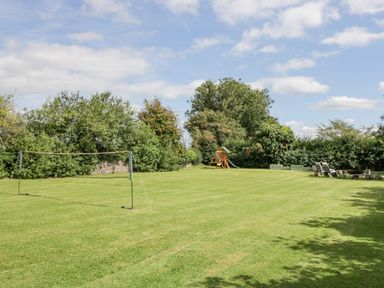A lawn with a badminton net a wooden playset and outdoor seating area at Orchard House Cottage in Malmesbury