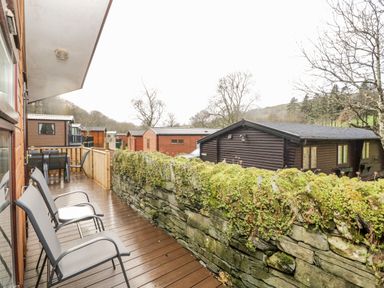 A balcony with chairs and a table overlooking wooden cabins and stone wall at Wansfell View in Limefitt Park near Troutbeck
