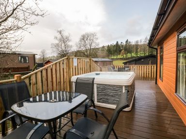 An outdoor wooden deck with a hot tub and a glass table with four chairs at Wansfell View in Limefitt Park near Troutbeck