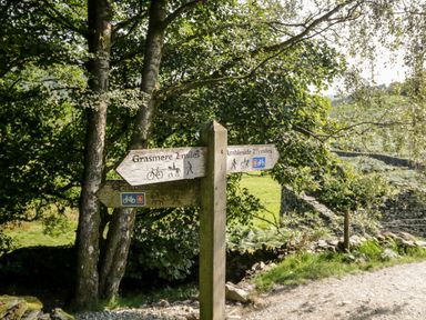 A wooden signpost on a gravel path showing directions to Grasmere and Ambleside near trees and a stone wall at Wansfell View Limefitt Park near Troutbeck