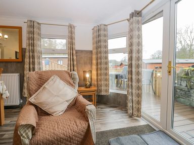 A living room with a chair covered by a quilted cover and cushion a side table with a lamp curtains on windows and glass doors at Wansfell View in Limefitt Park near Troutbeck