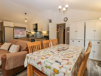 A dining area with wooden chairs and a table with a patterned tablecloth next to a kitchen with white cabinets and a brown sofa at Wansfell View in Limefitt Park near Troutbeck