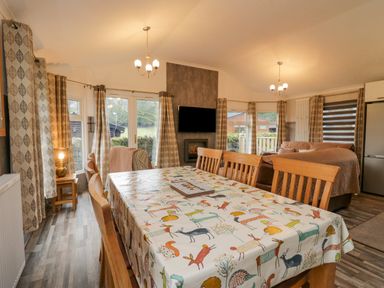 A dining area with a table covered with animal print tablecloth and chairs next to a living room with sofas and a wall-mounted TV at Wansfell View in Limefitt Park near Troutbeck