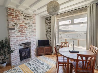 A dining area with a round wooden table and chairs near a window with a view of boats on the water and a stone fireplace with a small heater at Windycroft in Barmouth