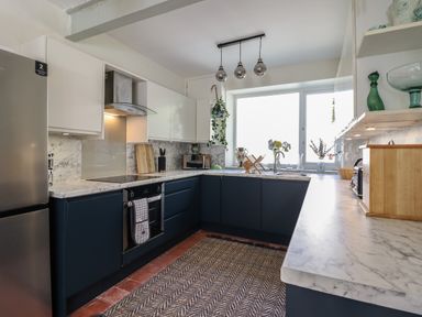 A kitchen with marble countertops dark lower cabinets white upper cabinets a stainless steel refrigerator oven and stove a window with plants and flowers at Windycroft in Barmouth