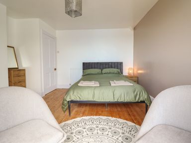 A bedroom with a green bedspread two beige chairs a patterned rug a wooden floor and a wooden chest of drawers with a mirror at Windycroft in Barmouth