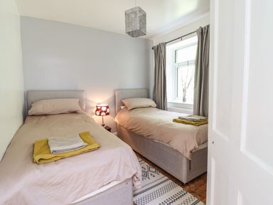 A bedroom with two single beds side by side each with beige bedding and folded towels a window with curtains and a bedside lamp between the beds at Windycroft in Barmouth