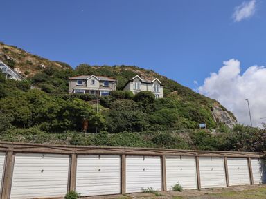A row of white garage doors in front of a hill with two houses and green vegetation under a blue sky at Windycroft in Barmouth