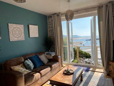 A living room with a brown leather sofa and cushions next to large open doors overlooking a beach and water at Windycroft in Barmouth