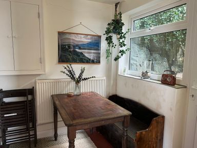A dining area with a wooden table, bench, folded chairs, a hanging plant, a window, and a framed picture at Windycroft in Barmouth
